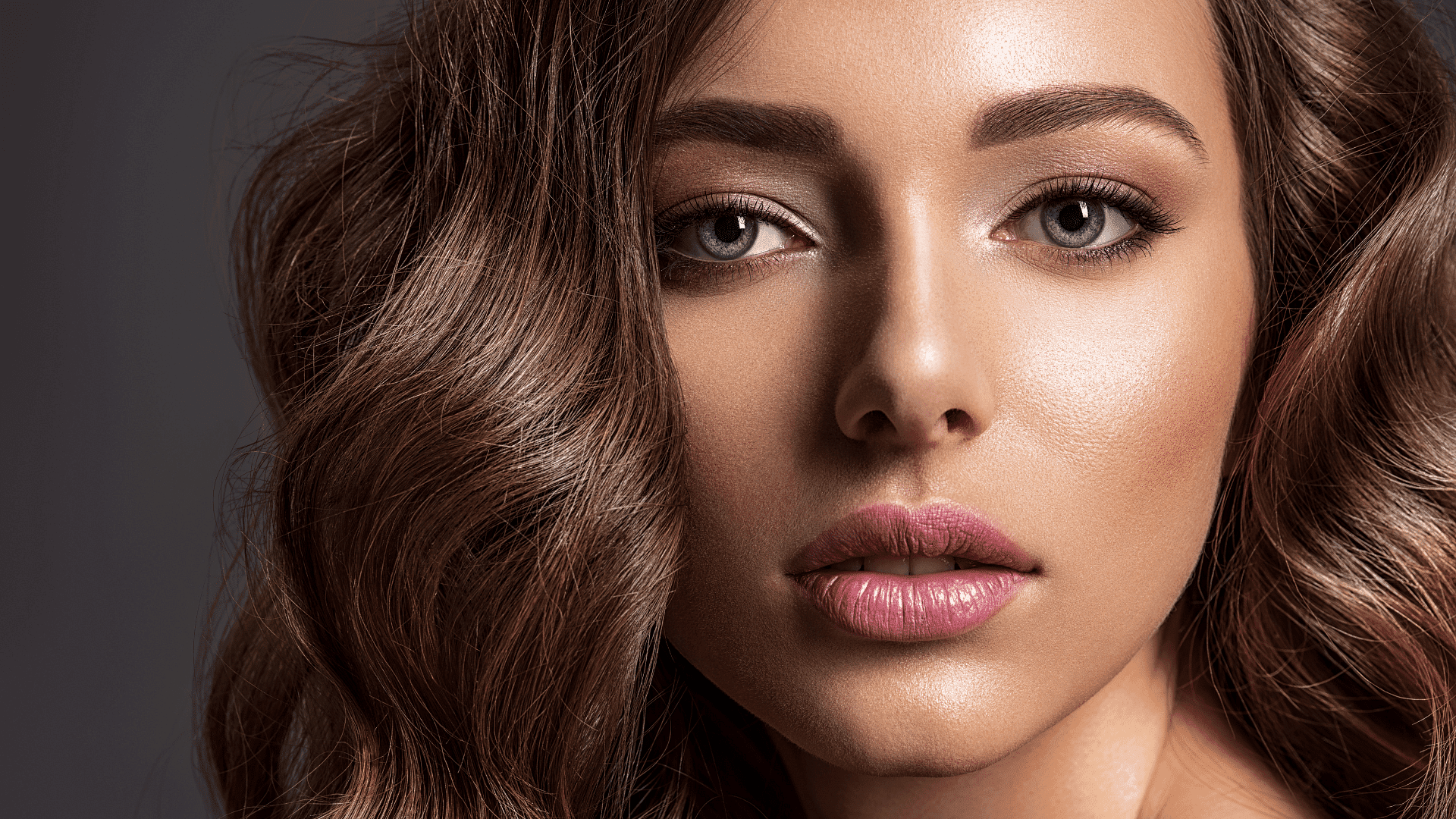 Close-up of a woman's face with wavy brown hair, pink lips, and a neutral expression.