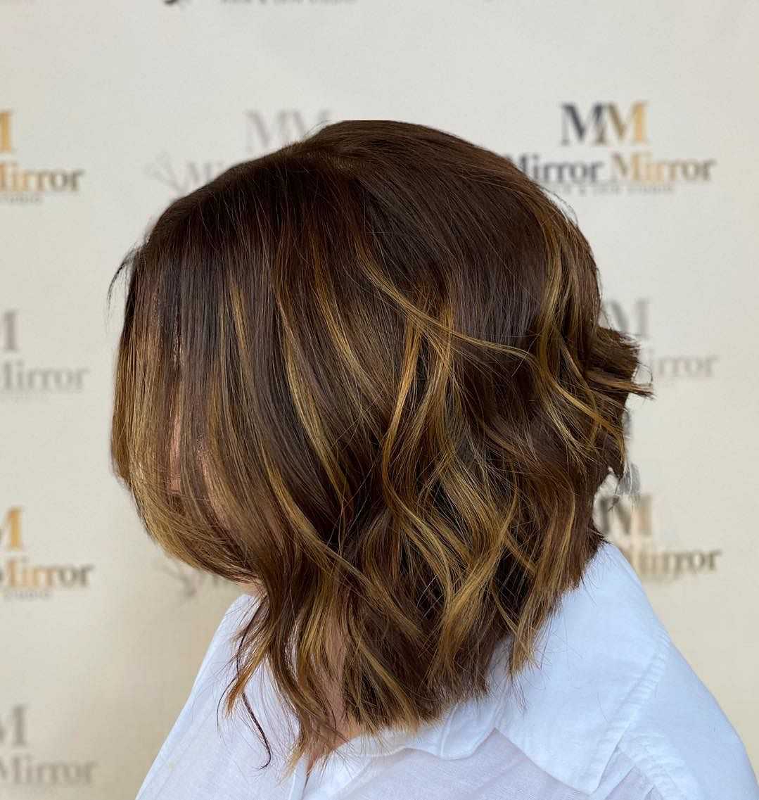 Woman with wavy, shoulder-length hair featuring brown and caramel highlights, in front of a salon backdrop.