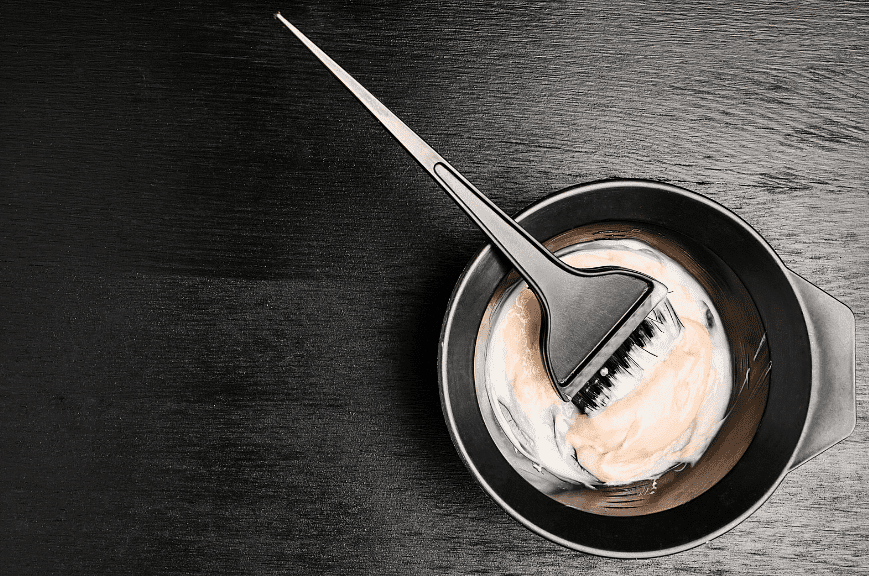 Bowl of hair dye with an applicator brush on a dark textured surface.