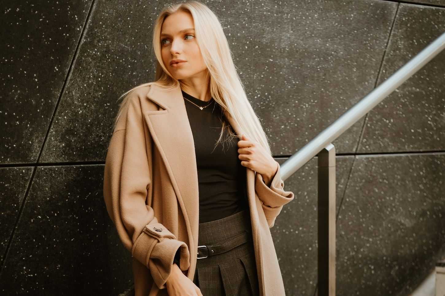 Young woman in a beige coat stands by a metal railing against a dark tiled wall.