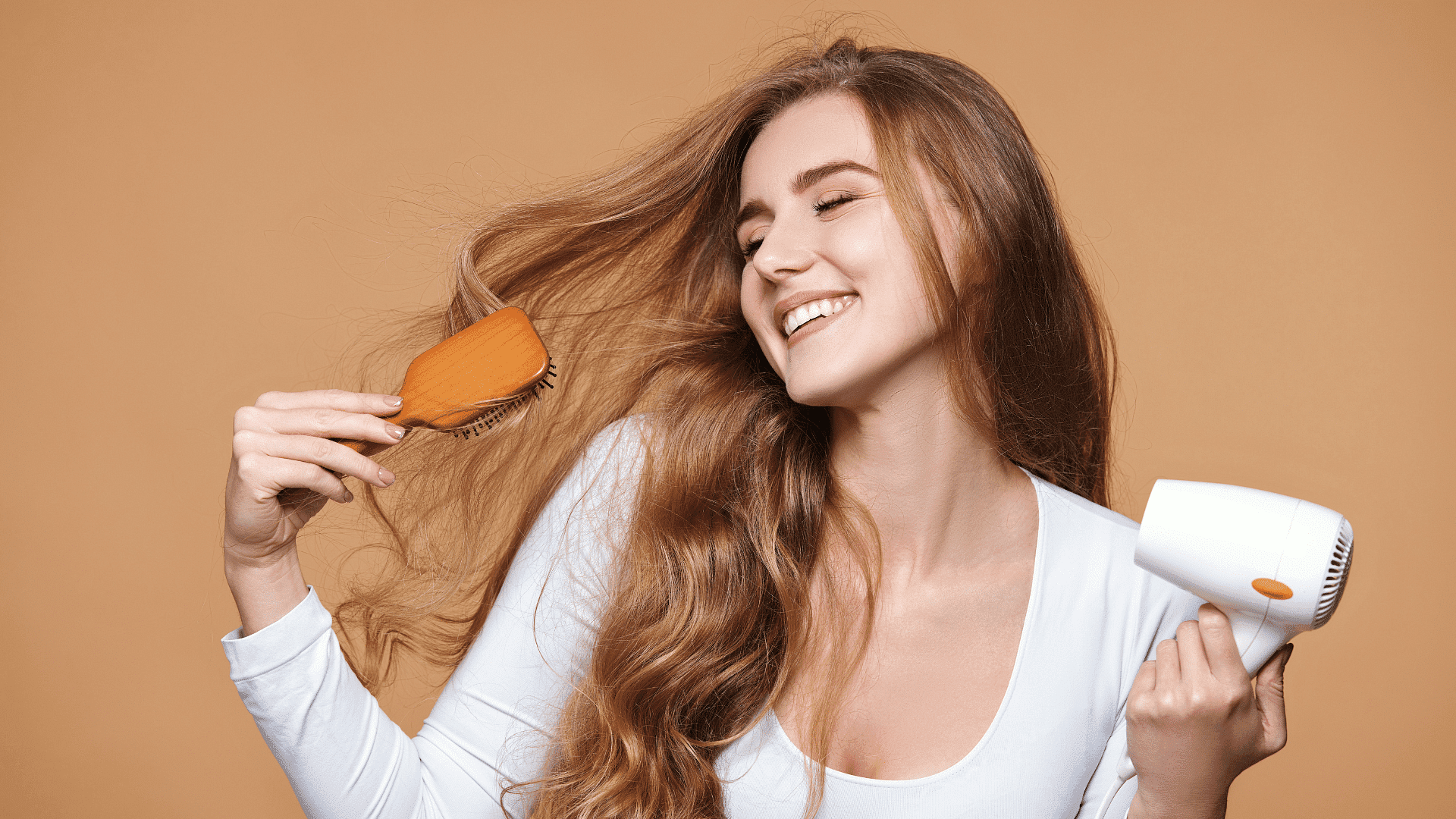 Woman smiling while brushing her long hair and holding a hairdryer against a tan background.