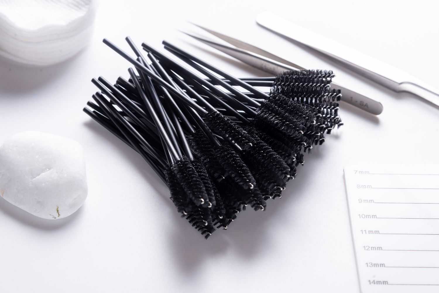 A pile of black mascara wands on a white surface with beauty tools in the background.